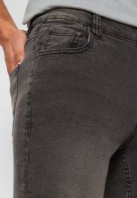 Jean en denim gris foncé avec une coupe slim, présentant un design classique à cinq poches et des variations de texture subtiles. Coutures renforcées en accents.