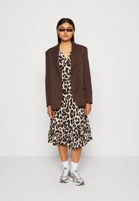 Brun blazer over en leopardmønstret kjole med volanger. Brukt med sølvfargede sneakers og hvite sokker. Bakgrunnen er helt hvit.