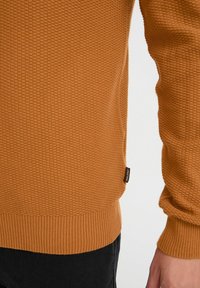 Waffle-textúrájú, égett narancssárga kötött pulóver, bordás szegéllyel és mandzsettával; oldalt logócímke található. Sötét nadrággal párosítva látható.