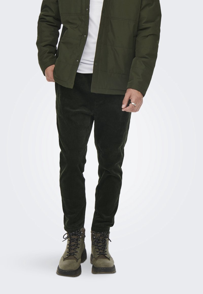 Groene pufferjack met drukknoopsluiting, gecombineerd met donkergroene ribfluwelen broeken en bruine veterschoenen met zwarte veters en rubberen zolen.