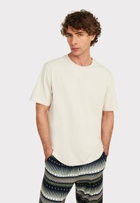 Cremfarbene Baumwoll-T-Shirt mit Rundhals und kurzen Ärmeln, kombiniert mit gemusterten Hosen mit grünen und blauen Zickzack-Mustern.