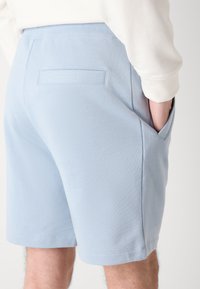 Șorturi de culoare bleu deschis, dintr-un material moale, având un buzunar pe spate și un brâu elastic, tăiate deasupra genunchiului, cu o textură netedă.