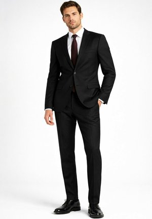 Homme debout, main dans la poche, portant un costume noir ajusté, une chemise blanche, une cravate foncée et des chaussures habillées noires brillantes, sur fond blanc.