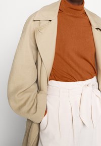 Manteau beige oversize, col roulé rouille, et pantalon blanc côtelé à taille haute avec ceinture nouée. Textures notables et détails de superposition.