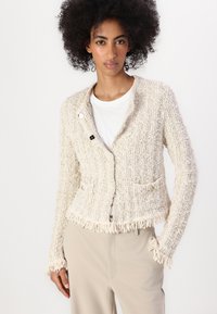 Cardigan in crema intrecciato con tessuto testurizzato, orlo con frange, due tasche frontali e bottoni decorativi. Abbinato a una camicia bianca e pantaloni chiari.