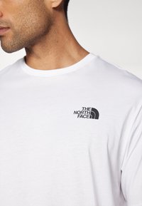 Camiseta blanca de cuello redondo hecha de suave algodón, con un pequeño logo negro en el pecho que presenta "The North Face" y un gráfico de media cúpula.
