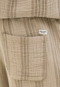 Beige színű pamut anyag vízszintes barna csíkokkal és zsebbel. Jellemzője a "JACK & JONES EST. 1990." feliratú címke. Texturált felszín.