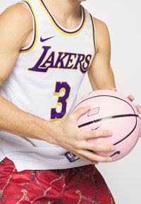 Maglia da basket bianca con testo "Lakers" in viola e numero 3, abbinata a pantaloni corti rossi in rete con un motivo a catena, mentre tiene un pallone da basket rosa.