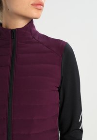 Primo piano di una persona che indossa un gilet trapuntato viola senza maniche sopra una maglietta nera a maniche lunghe con zip.
