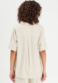 Chemise en lin beige à manches courtes, avec col et pli au dos. Texture douce avec une coupe décontractée et un ourlet arrondi.