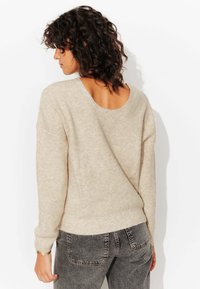 Pull en maille côtelée beige avec décolleté large et dos ouvert, associé à un jean gris foncé avec poches arrière et coupe ajustée.