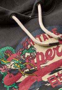 Sudadera negra con un gráfico de dragón vintage colorido con acentos en verde, amarillo y rojo. Material de algodón con cordones blancos y herrajes metálicos.