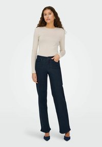 Lys beige langermet topp med bølget halslinning, kombinert med mørkeblå vide jeans. Modellen står i blå hæler, med hendene i lommene.