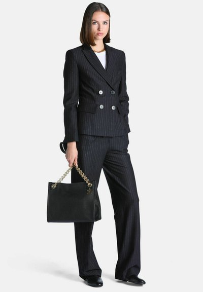 Femme portant un costume noir à fines rayures et double boutonnage, tenant un sac à main noir avec une bandoulière chaîne dorée.