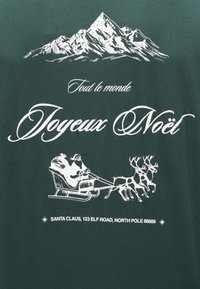 Camiseta verde con estampado blanco de montañas, texto "Tout le monde Joyeux Noël" y Santa Claus con renos en un trineo.
