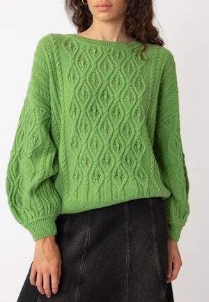 Grüner gestrickter Pullover mit einem strukturierten Rautenmuster, weiten Ärmeln und ribbelt Saum, kombiniert mit einem dunklen Jeansrock.