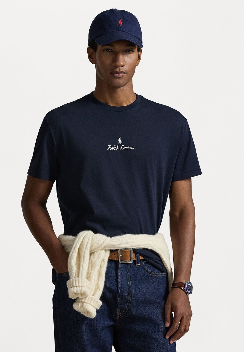 T-shirt de algodão azul-marinho com o logótipo branco "Ralph Lauren", combinado com um suéter de malha em creme claro atado à cintura e jeans de ganga escura.