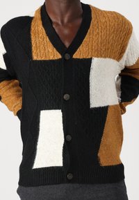 Sweter rozpinany z dzianiny z blokami kolorów czarnego, białego i musztardowego, teksturowanymi wzorami oraz czarnymi, okrągłymi guzikami na przodzie.