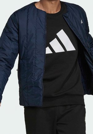 Mann trägt eine marineblaue Jacke mit Reißverschluss über einem schwarzen Adidas-Sweatshirt mit großem weißem Adidas-Logo auf der Brust.