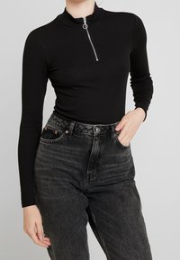 Svart ribbad långärmad tröja med dragkedja och ståkrage, tillsammans med högmidjade svarta denimjeans med kopparnitar och fickor.