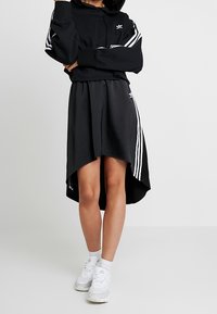 Sudadera corta negra con tres rayas blancas en las mangas, combinada con una falda asimétrica negra con rayas blancas. Lleva zapatillas blancas.