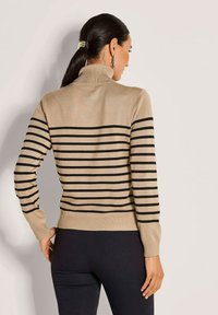 Frau mit dunklen Haaren in einem Pferdeschwanz trägt einen beigen Rollkragenpullover mit schwarzen horizontalen Streifen und schwarzen engen Hosen und schaut von hinten.