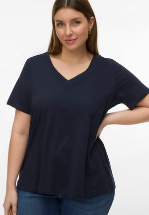 Woman with straight brown hair wearing gold hoop earrings and a navy blue V-neck short sleeve shirt with blue jeans, posing with one hand on hip.