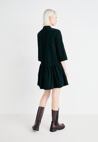 Robe en velours côtelé vert foncé avec une jupe à volants et des manches trois-quarts, associée à des bottes en cuir marron jusqu'aux genoux avec des accents noirs.