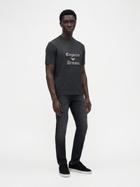 Άνδρας στέκεται φορώντας μαύρο T-shirt Emporio Armani, σκούρα γκρι στενά τζιν και μαύρα αθλητικά παπούτσια με λευκές σόλες πάνω σε λευκό φόντο.