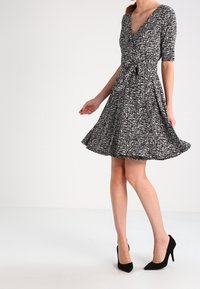 Svart och vit mönstrad omslagsklänning med korta ärmar, figursydd midja, utsvängd kjol och slät textur. Bärs med svarta högklackade skor.