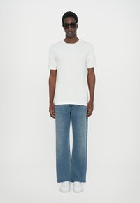 Hvit t-skjorte med rund hals, blå vide jeans og hvite joggesko. Modellen har på seg mørke solbriller, med en enkel bakgrunn.