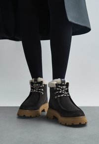 Donkerbruin suède enkellaarzen met crèmekleurige nep bontafwerking, zwarte veters en een chunky beige zool. Logo-detail op de tong.