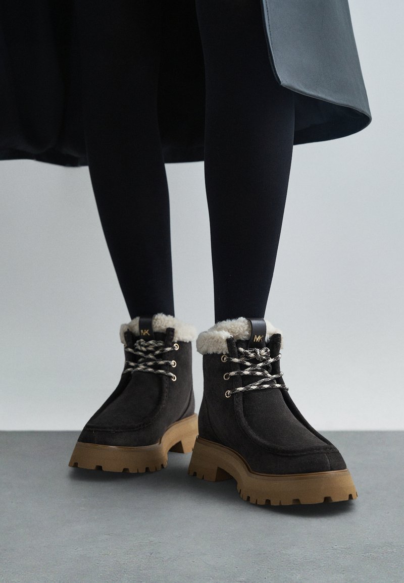 Donkerbruin suède enkellaarzen met crèmekleurige nep bontafwerking, zwarte veters en een chunky beige zool. Logo-detail op de tong.