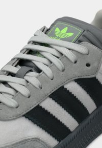 Γκρι σουέντ αθλητικό παπούτσι με τρεις μαύρες ρίγες, χοντρές γκρι κορδόνια και φωτεινή πράσινη λογότυπη επιγραφή στη γλώσσα. Διαθέτει υφασμένη μαύρη καουτσούκ σόλα.