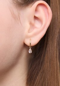Złota kolczyk w kształcie obręczy z kroplą przezroczystego kryształowego wisiorka, prezentowany na uchu modelki. Projekt podkreśla prostotę i elegancję.