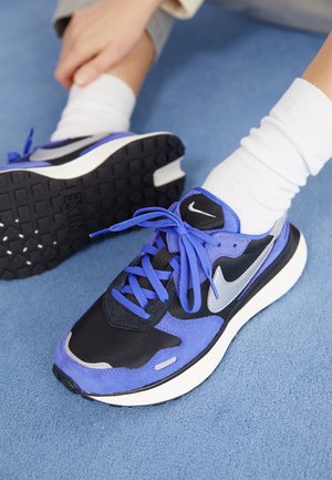 Osoba nosząca białe skarpetki oraz niebiesko-czarne sneakersy Nike siedzi na niebieskim dywanie, poprawiając but na jednej stopie.