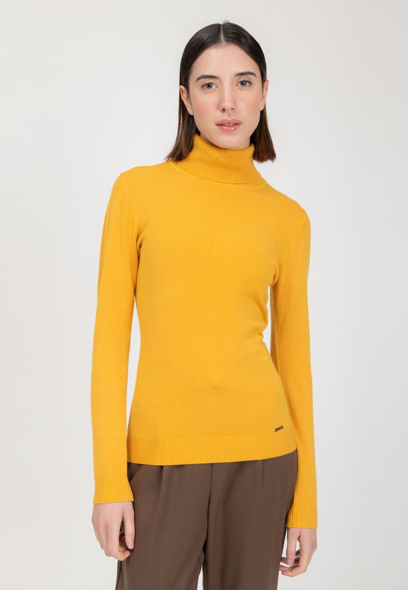 Langärmliger, enganliegender Rollkragenpullover aus leuchtend gelbem Strick, mit gerippten Bündchen und Saum, kombiniert mit braunen Hosen.