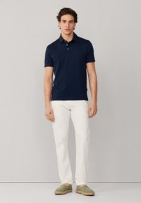 Hombre de pie contra una pared lisa, vestido con un polo azul marino de manga corta, pantalones blanco roto y zapatos verdes tipo slip-on.