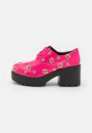 Heller pinker Plateauschuh mit klobigem schwarzem Absatz, blumenförmigen Strassverzierungen und Schnürdesign auf weißem Hintergrund.
