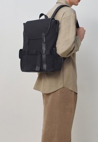 Zaino in tessuto nero con dettagli in pelle, numerose tasche, spallacci regolabili e una finitura testurizzata distintiva. L'utente indossa una maglietta beige.