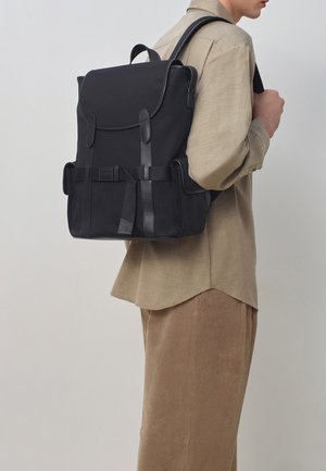 Schwarzer Stoff-Rucksack mit Lederelementen, mehreren Fächern, verstellbaren Riemen und einer charakteristischen strukturierten Oberfläche. Der Benutzer trägt ein beiges Shirt.
