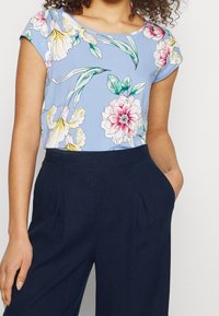 Blusa floreale di colore blu chiaro con maniche corte e scollatura rotonda, caratterizzata da motivi di fiori rosa e gialli, abbinata a pantaloni blu navy scuro.