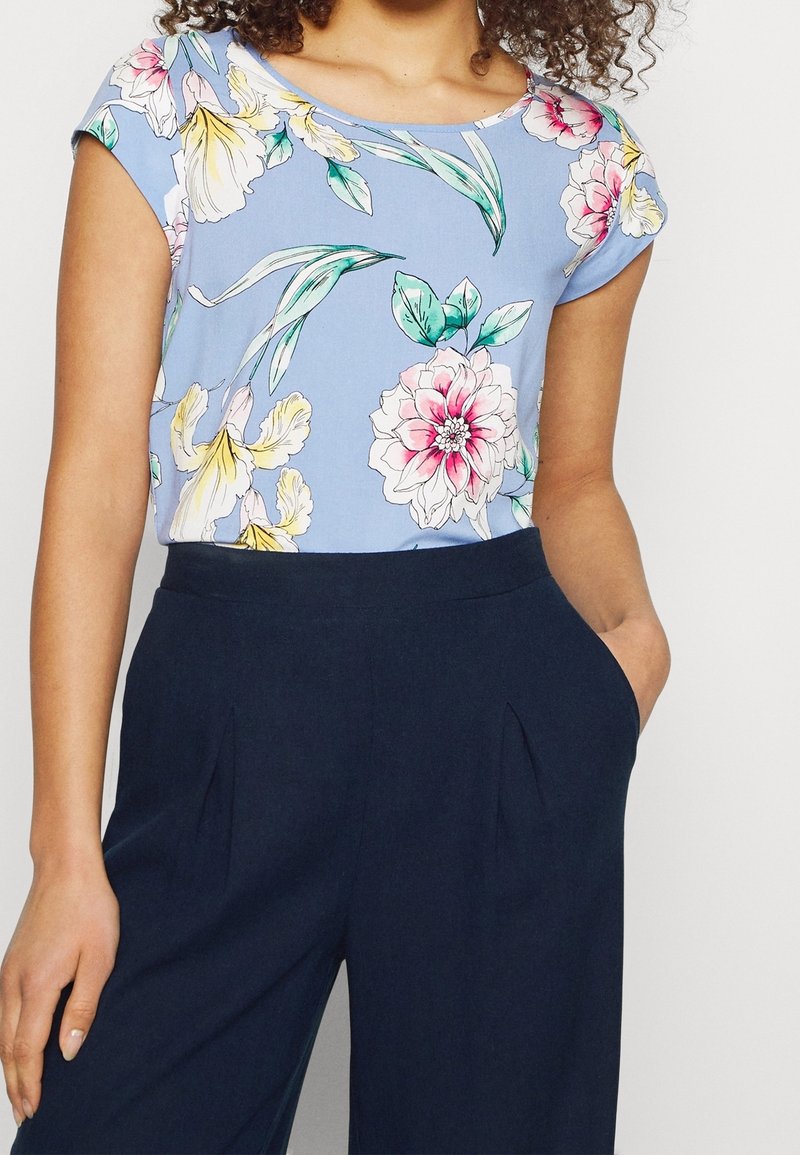 Blusa floreale di colore blu chiaro con maniche corte e scollatura rotonda, caratterizzata da motivi di fiori rosa e gialli, abbinata a pantaloni blu navy scuro.