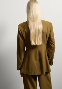 Chaqueta marrón ajustada con una silueta estructurada, hombros marcados y mangas largas, combinada con pantalones de talle alto a juego.