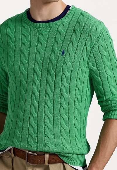 Pulover verde tricotat cu un model în cablu, gât rotund și manșete cu canal. Prezintă un mic logo albastru pe piept. Combinat cu pantaloni khaki.