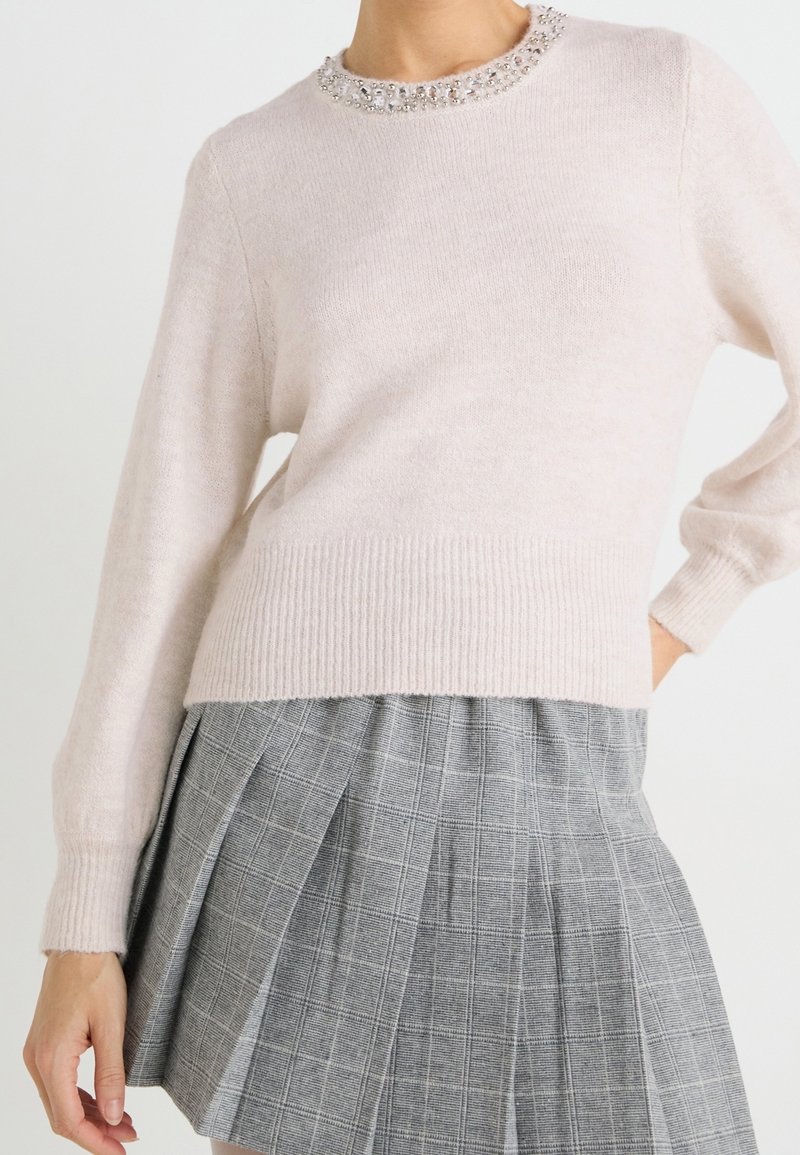 Femme portant un pull rose clair avec un col orné de bijoux et une jupe plissée grise à taille haute avec un motif à carreaux.