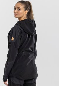 Black waterproof jacket with a hood, featuring a textured material, elastic cuffs, and a subtle leather patch on the shoulder.