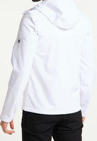 Biała kurtka z kapturem z pikowanymi łatami na ramionach i łokciach, noszona przez osobę zwróconą tyłem, w połączeniu z czarnymi spodniami.