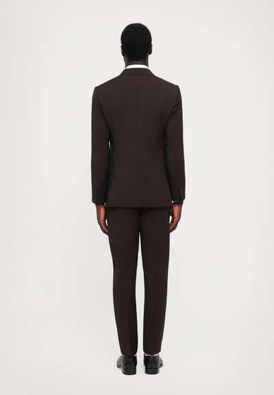 Abito marrone scuro con una vestibilità su misura, caratterizzato da un unico spacco e una sottile trama. Scarpe nere completano il look. Vista da dietro.