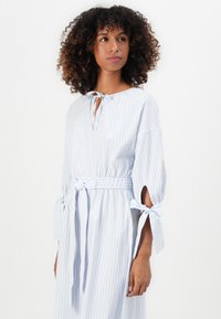 Mulher com cabelo encaracolado vestindo um vestido listrado em azul claro e branco, com laços nas mangas e na cintura, olhando ligeiramente para a sua esquerda.